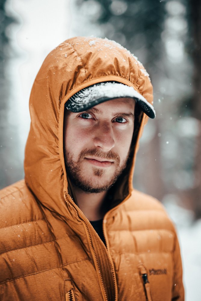A man wearing a orange winter jacket and a cap, with snow on his jacket and cap, standing outdoors in a snowy environment.