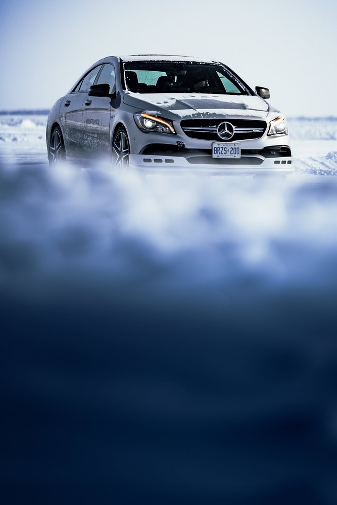 A silver Mercedes-Benz sedan parked on snowy ground with snow on its hood and roof, under a cloudy sky.