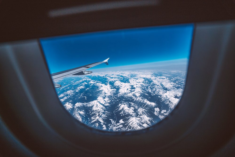 View of snow-capped mountain peaks seen from an airplane window with part of the airplane wing visible.