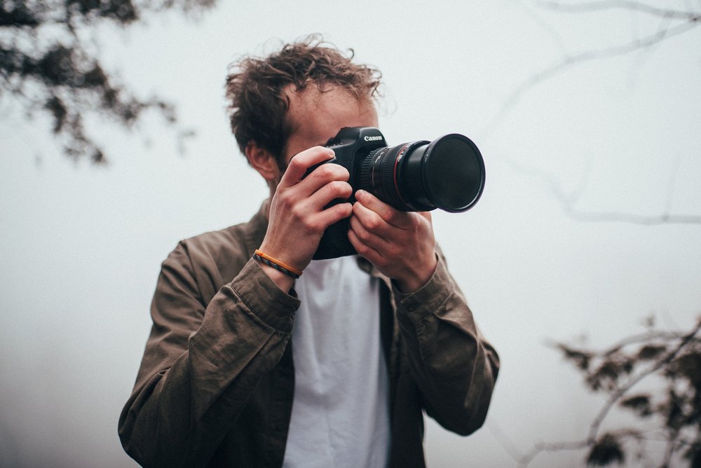A young man with curly hair wearing a brown jacket and bracelet, holding a Canon camera up to his face, outdoors against a cloudy sky with tree branches.