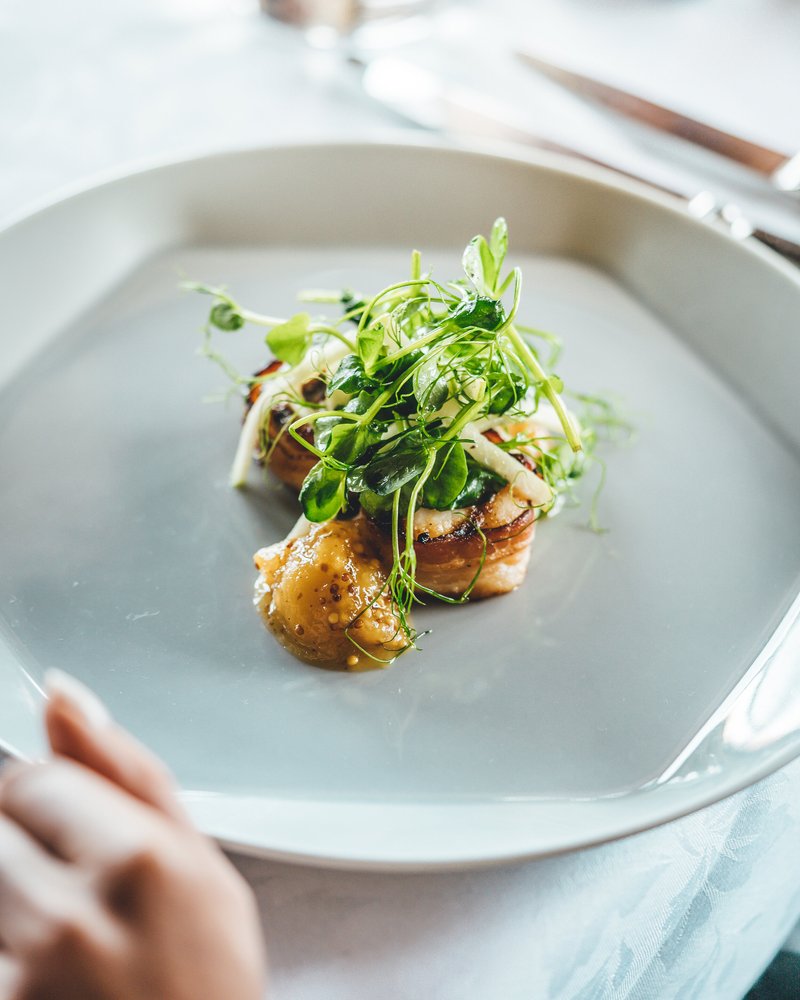 Close-up of a plated gourmet dish with microgreens on top, served on a white plate with a blurred background.