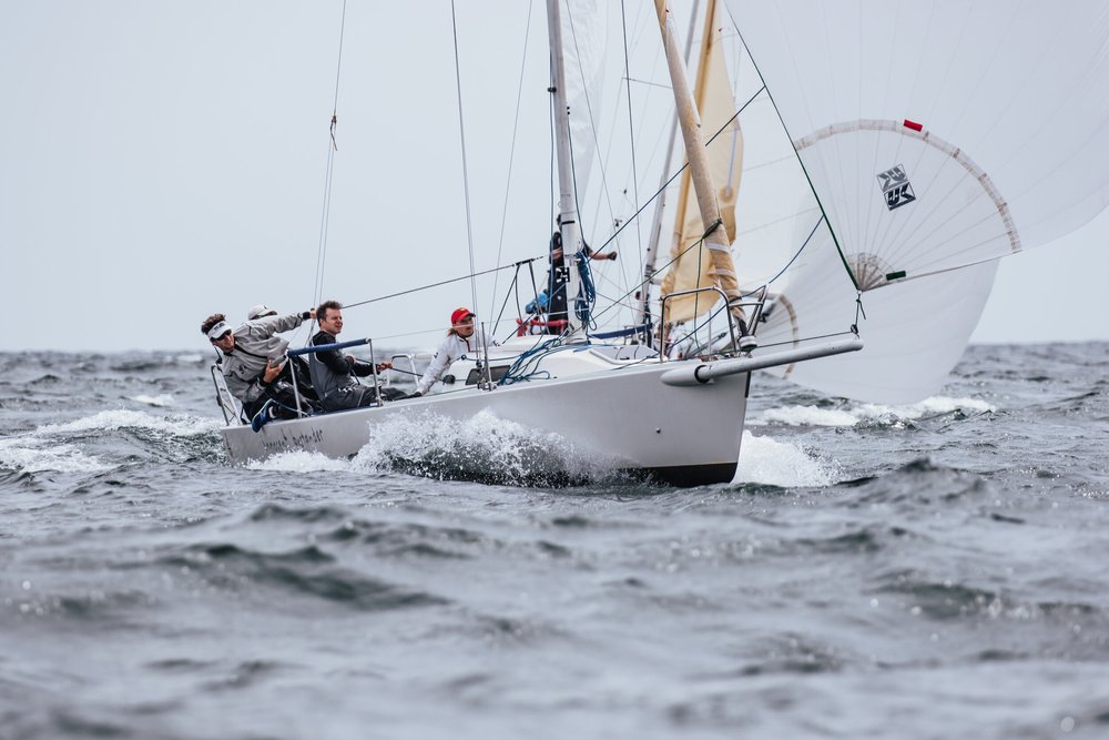 A sailboat with four people onboard navigating through choppy waters, with the sail fully deployed and the boat tilted to one side.