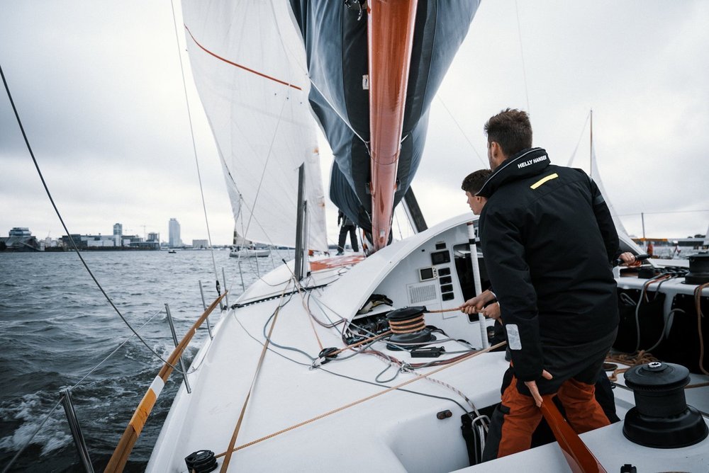 Crew members working on a sailboat during daytime, with city skyline and cloudy sky in the background.