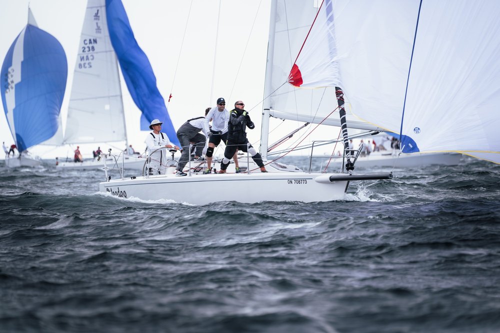 A sailboat with four crew members navigating through choppy water on a cloudy day, with other sailboats visible in the background.