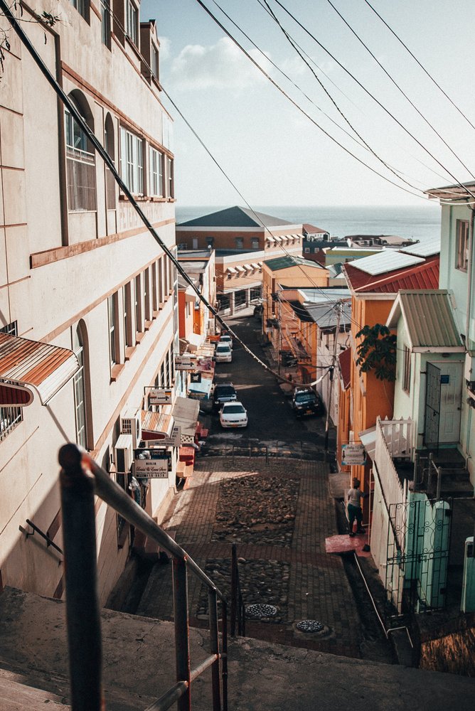 A street view showing multi-story buildings, parked cars, and overhead power lines with the sea in the background on a sunny day.