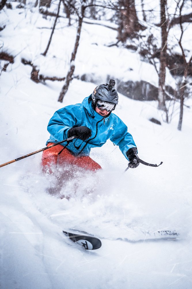 A person wearing a helmet, goggles, and a blue jacket skiing downhill on a snowy slope, with snow flying up around him and snow-covered trees in the background.