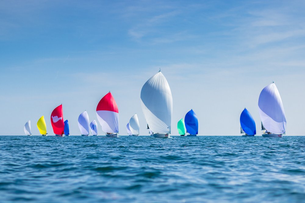 Multiple sailboats with colorful spinnakers on a calm ocean under a clear blue sky.