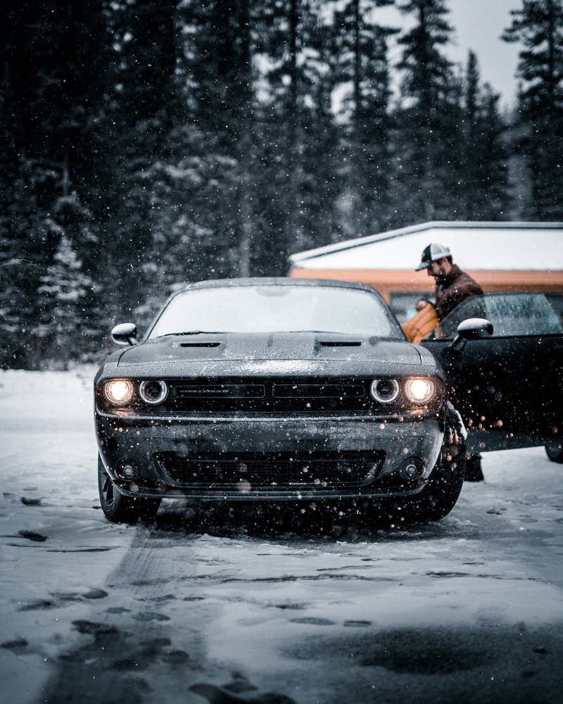 A black sports car is parked on a snow-covered surface, with snow falling around it. In the background, there are tall trees and a small building, possibly a garage or shed. A person wearing a helmet and brown clothing is standing near the car, possibly preparing to get in or out.