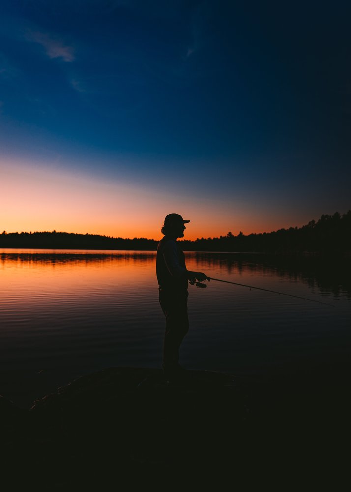 Silhouette of a person fishing at sunset by a lake, with a colorful sky transitioning from orange to deep blue, and trees in the background.