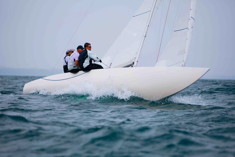 Three people sailing on a white sailboat on a calm body of water with a cloudy sky overhead.