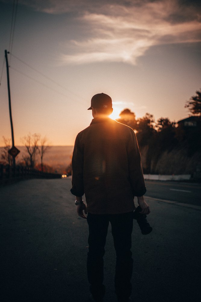A person walking on a road during sunset, carrying a helmet and a backpack, with trees and power lines in the background.